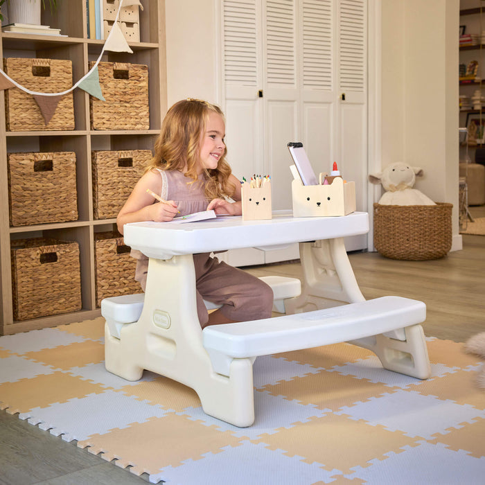 Girl with long blonde hair sitting at the table with pen and paper. Setting is a playroom with shelves and table is sitting on play mats.