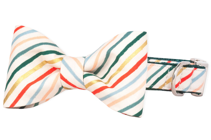 Dog collar with an attached matching bow tie in a festive stripe pattern. The cream background is decorated with diagonal stripes in red, green, metallic gold, light blue, and blush, giving it a candy cane–inspired holiday look.