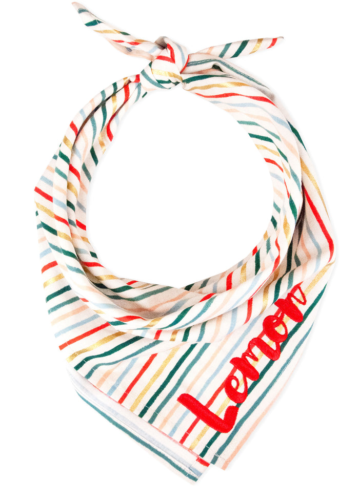 A holiday bandana for dogs, designed with diagonal stripes in vibrant seasonal colors: red, green, metallic gold, pastel blue, and peach. The cream background highlights the playful, festive design. The name "Lemon" is embroidered on the bottom right.