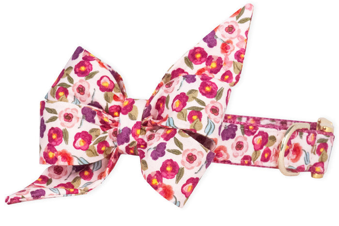 Dog collar with an oversized belle bow made from fabric patterned with vivid small flowers in shades of pink, accented with leaves on a pale pink background. The belle bow features two tall pointed loops and wide, layered lower folds for a feminine look. Both bow and collar are made in the same fabric.