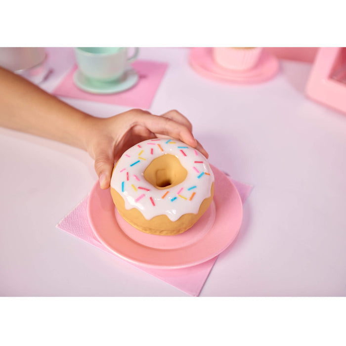 A hand holds a donut-shaped object with white frosting and multicolored sprinkles on a pink plate, set on a pastel pink table with cups in the background.