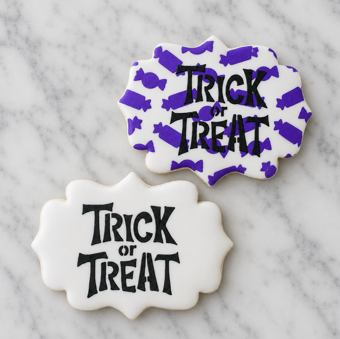 Two white, plaque shaped Halloween sugar cookies decorated with the Tricky Trick or Treat Cookie Stencil in black royal icing and one of the cookies also is stenciled with the Candy Pattern in purple. Both cookies shown on a Carrara Marble counter, BakeArt Stencils